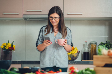 Happy Woman Using Mobile Phone while Drinking Cup of Tea in the Kitchen. 
Smiling female chef holding a cup of coffee and typing text message on her smartphone or reading online recipe at home.