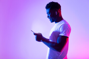 Online life concept. Millennial black guy using mobile phone, checking web or social media in neon light, copy space - obrazy, fototapety, plakaty