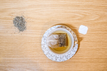 Vista cenital de una taza de té con una bolsita de té de manzanilla en el interior. Hierbas secas en la mesa. Adornos florales dorados. Fondo de madera