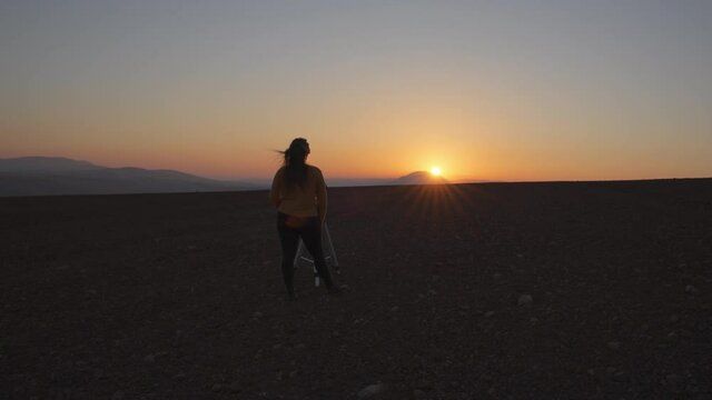 Wide angle view of a woman filming a sunset with a camera and a tripod in the desert. Slow motion. 