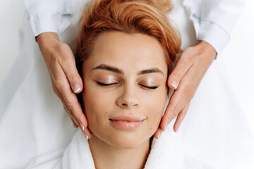 Obraz na płótnie Canvas Top view of relaxed woman laying with closed eyes during anti-aging facial treatments while cosmetologist massaging her face with hands