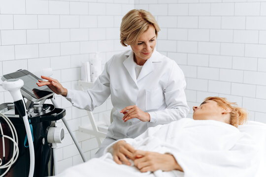 Relaxed woman lying on couch while cosmetologist adjusting settings on monitor for laser hair removal device. Stock photo