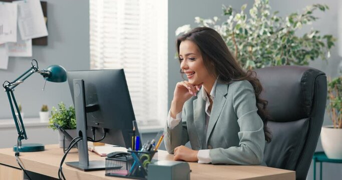 Positive woman with long hair, wearing a blazer, sits at her desk in office in front of a computer screen, has written off her last email, smiles in a break from work contentedly dances joy