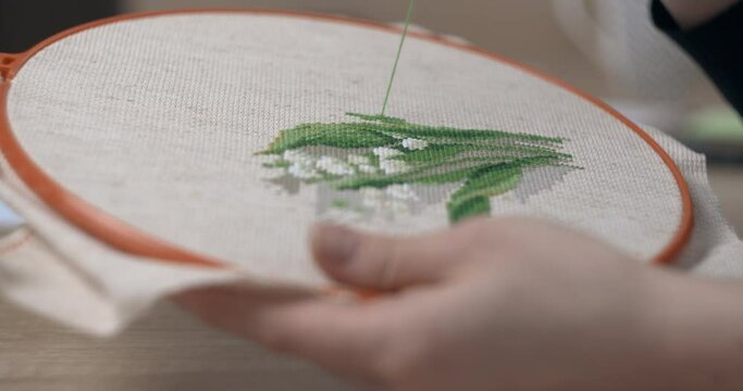 A woman is embroidering lilies of the valley with a cross on a canvas fixed with a hoop. Close-up