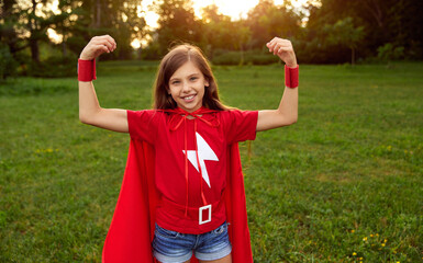 Playful girl in superhero cape smiling and showing muscles in park