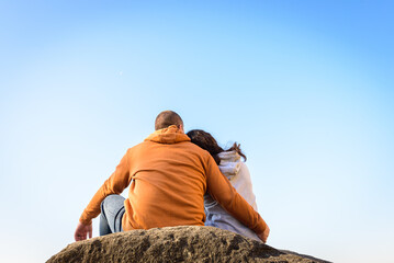 young couple hugging and sitting close, back, on a large rock in the background of the lake