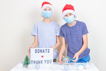 Teenager boys volunteers with protective face mask preparing to collect Christmas donations indoors. Kids standing near desk with Christmas decorations and lightbox with message Donate and Thank you