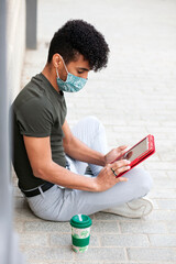 Young African American student with face mask, concentrating on using red tablet while sitting on the street with his coffee.