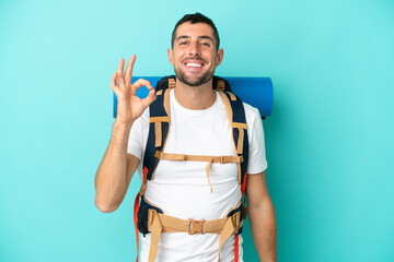 Young mountaineer caucasian man with a big backpack isolated on blue background showing ok sign with fingers