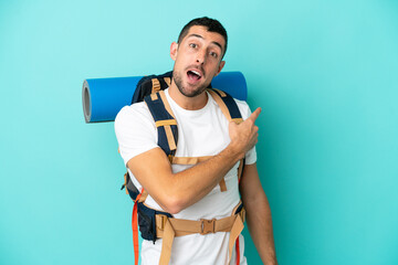 Young mountaineer caucasian man with a big backpack isolated on blue background surprised and pointing side