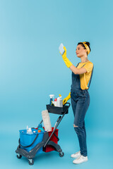 cleaner in denim overalls and rubber gloves taking selfie near cart with cleaning supplies on blue.