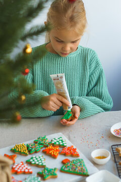 A cute 8-year-old girl in a light green sweater paints a festive Christmas, New Year cookie in the form of a Christmas tree. Bright festive Christmas, New Year cookies prepared at home, with your own 