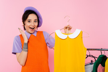 amazed woman showing wow gesture near yellow sleeveless dress and rack with clothes isolated on...