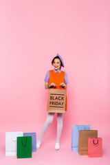 cheerful woman holding shopping bag with black friday lettering near purchases on pink.
