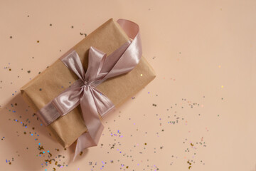Obraz premium Birthday or New Year's eve present in kraft paper with pink ribbon on a soft beige background with glitter and confetti. Xmas composition. Flat lay. Happy holidays celebration and giving love concept
