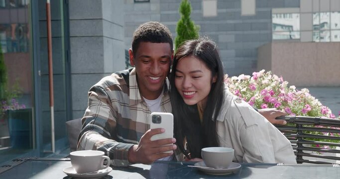 Young multiracial loving couple sitting together in the outdoor cafe on beautiful sunny day at romantic date , taking a self portrait and posting image on social media.