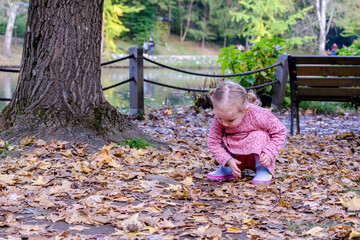 Blonde little girl wear pink dress and rain boots and crouch on the ground and looking for something , autumn concept idea