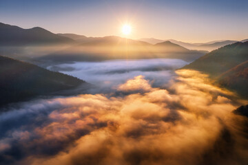 Fototapeta na wymiar Mountains in low clouds at sunrise in autumn. Aerial view of mountain peak in fog in fall. Beautiful landscape with rocks, forest, sun, colorful sky. Top view of mountain valley in clouds. Foggy hills