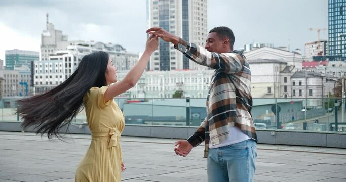 Beautiful multicultural loving couple of two cheerful people dancing at romantic date on the rooftop of an urban building. Handsome African guy turning his girlfriend around herself while dancing