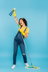 excited woman in rubber gloves and denim overalls singing with detergent and mop on blue.
