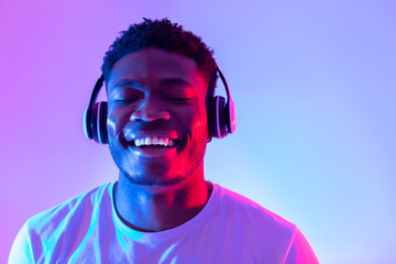 Attractive young black guy in casual wear listening to music in headphones, closing eyes and relaxing in neon light