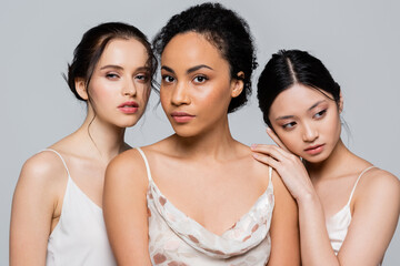 Multicultural women in satin dresses posing isolated on grey