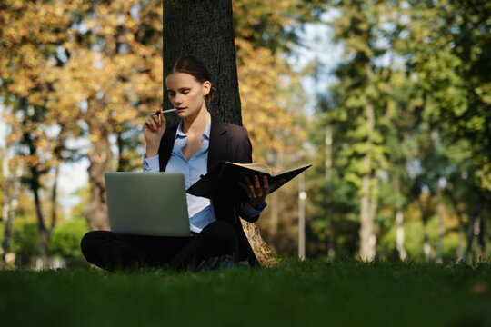 Side view of woman sitting in a park on green grass with a lapot and notebook, outdoor worker, remote work.
