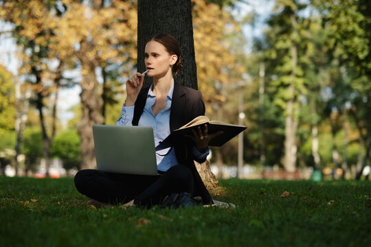Side view of woman sitting in a park on green grass with a lapot and notebook, outdoor worker, remote work.