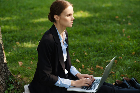 Side view of woman sitting in a park on green grass with a lapot and notebook, outdoor worker, remote work.