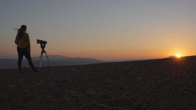 Low angle dolly in view of a woman filming the sunset with camera and tripod in the desert. Slow motion.