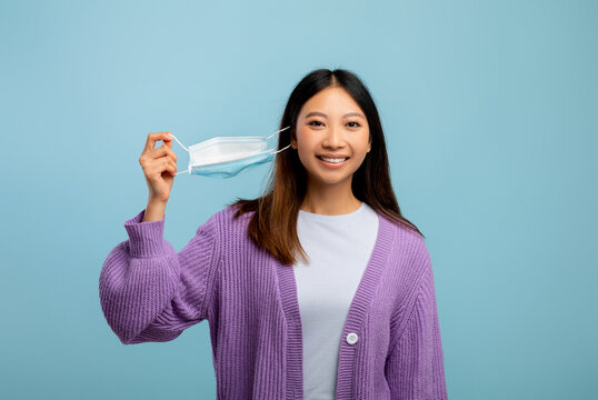 Young asian woman taking off disposable protective face mask, standing over blue studio background, breathing fresh air