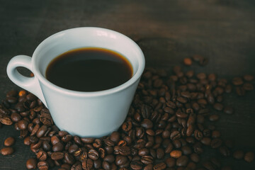 White cup filled with coffee, surrounded by coffee beans on a dark wooden background. Copy space. Pure black coffee