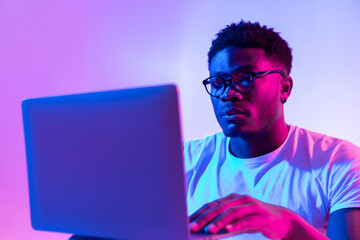 Focused young black man in glasses using laptop computer for online work or communication in neon...
