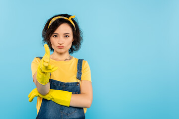 woman in denim overalls and rubber gloves showing gun gesture isolated on blue.