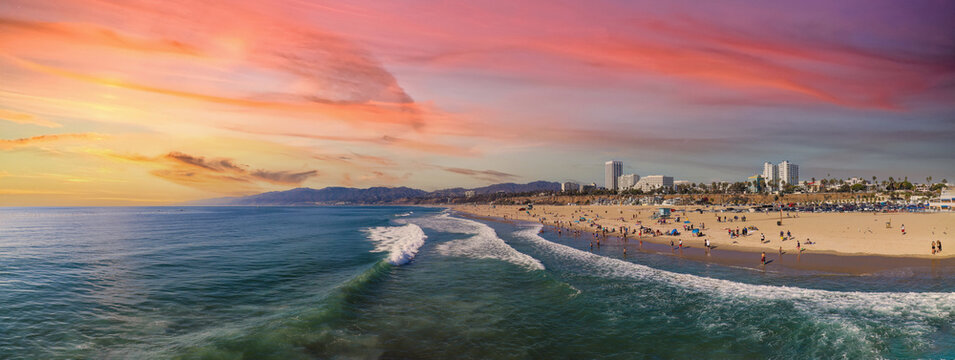 a stunning panoramic shot of large waves form the vast blue ocean rolling into the beach filled with people relaxing with colorful umbrellas at sunset with powerful clouds at Santa Monica Beach