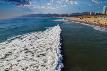 Fototapeta na wymiar a majestic shot of the large waves breaking in vast blue ocean water with a sandy beach filled with people and the cityscape with buildings in the background, blue sky, mountain ranges in California