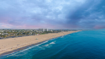 Fototapeta na wymiar an aerial shot of the Santa Monica beach with people relaxing on the beach near the blue ocean water with color umbrellas, the cityscape with buildings and trees with powerful clouds in California US