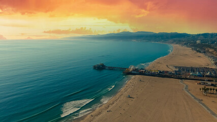 an aerial shot of the beach with silky brown sand, vast blue ocean water, palm trees, hotels along the beach, a bike path and mountain ranges, a pier at sunset at Santa Monica Beach in California