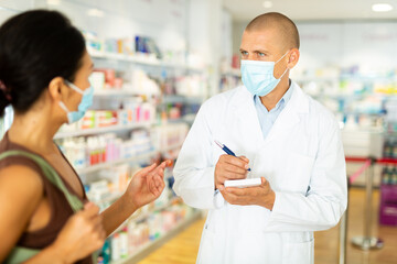 Pharmacist in protective mask writes in notebook the name of the drugs that the buyer tells him to