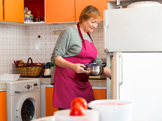 Smiling aged housewife putting pan in fridge in cozy home kitchen..