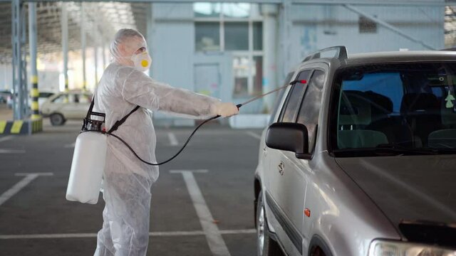 Man dressed white protective overalls spraying vehicle antibacterial sanitizer sprayer quarantine Caucasian person protective suit disinfects car on parking during covid pandemic New normal prevention
