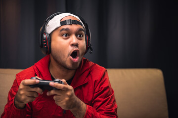 Excited and shocked face of Asian gamer with headphone holding headset playing video game online sitting on sofa at living room. Indian professional gamer streaming on social playing game very fun © Chanakon