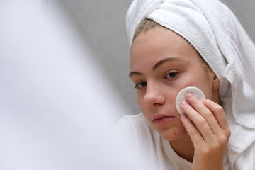 A teenage girl wiping her face with a cotton pad near the mirror. Facial skin care in adolescence.