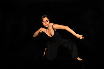 Full body portrait of a latin woman posing with one knee on the ground and moving her arms isolated on black background. Female body expression concept.