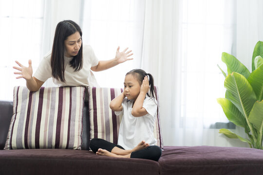 Asian little girl covering ears while mother scolding her in living room - Powered by Adobe