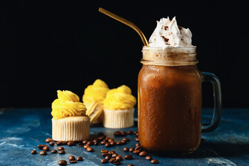 Mason jar of tasty iced coffee with cream and cupcakes on table