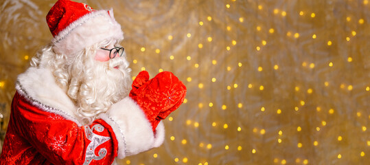 Portrait of santa clois with glasses blowing on mittens against the background of garlands. Selective focus. Beautiful christmas card