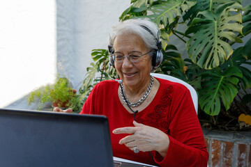 Happy grandmother looks at laptop camera, wears headphones, talks to family or communicates with...
