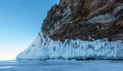 The base of the granite rock, devoid of vegetation, is covered with a thick layer of bizarre icicles.  The texture of the stone. Fragments of ice on the surface of a frozen lake. Blue sky. Baikal