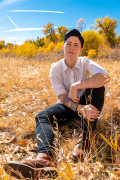 Woman sitting in a meadow during autumn with leaves and several trees. The woman has short hair and looks serious